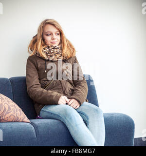 Schöne blonde kaukasische Teenager-Mädchen in warme Kleidung über weiße Wand Hintergrund auf blauem Sofa sitzen Stockfoto