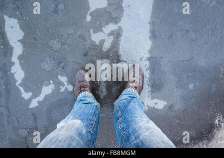 Männliche Füße stehen auf gefrorenen Pfütze mit dünnem Eis und fallende Blätter Stockfoto