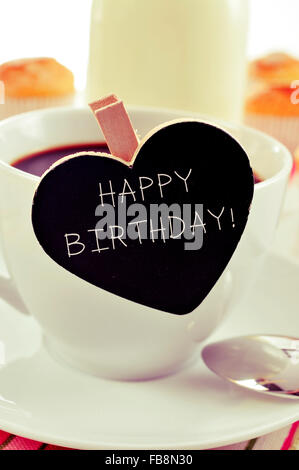 der Text happy Birthday geschrieben in einer Herzform Tafel platziert in einer Tasse Kaffee, mit einigen Muffins im Hintergrund in einer Stockfoto