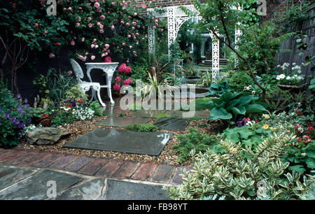 Weißen Schmiedeeisen Tisch und Stuhl auf der Terrasse im Reihenhaus Garten mit rosa Rosen und einen Spiegel an der Wand unter weißen Pavillon Stockfoto