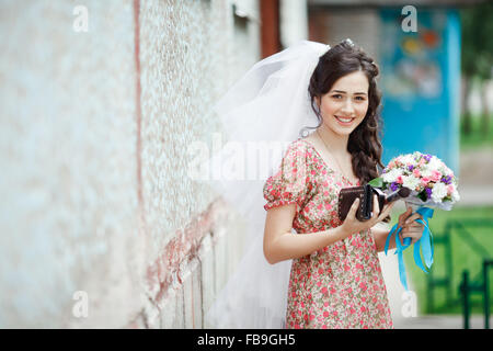 Die Braut in einem schlichten Retro-Kleid mit Blumenmuster, bereits tragen Schleier, Brautstrauß und Handtasche, posiert vor Haus, direkt in die Kamera, lächelnd. Stockfoto