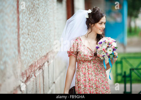 Die Braut in einfachen Retro-Kleid mit Blumenmuster, bereits tragen Schleier, Brautstrauß und Handtasche, posiert vor Haus, auf der Suche zur Seite. Stockfoto