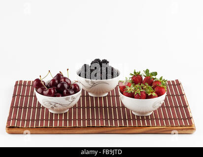 Drei Keramikschalen mit Beeren - Erdbeeren, Brombeeren und Kirschen auf Bambusmatte isoliert auf weiss Stockfoto