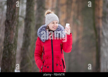 Glückliches schöne Mädchen Strickmütze und roten Wintermantel, im Freien, mit Handy, sieht auf dem Bildschirm des Smartphones Stockfoto