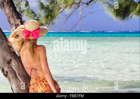 Junge Frau in einem Sarong und Hut suchen heraus zum Meer und gegen einen Baum, Rum Point, Grand Cayman-Inseln Stockfoto