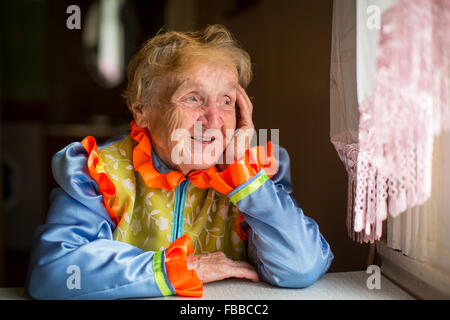 Alte Frau in traditioneller Tracht sitzt am Fenster im Haus. Stockfoto