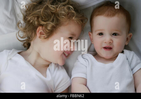 Querformat Hochformat eines lächelnden blonden Kleinkind Jungen und seinen roten Haaren Babyschwester, beide tragen weiße Unterhemden Verlegung auf einem weißen Blatt. Stockfoto