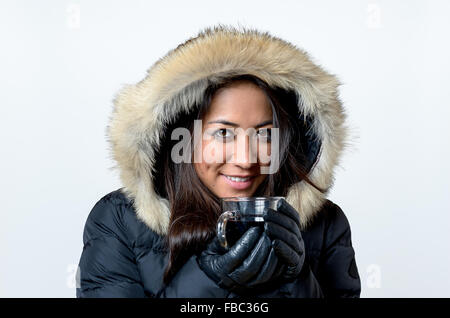 Lächelnde junge Frau einem heißen Getränk die Becher in ihre behandschuhten Hände umklammern, wie sie sich im warmen pelzigen Kapuze, k kuschelt Stockfoto