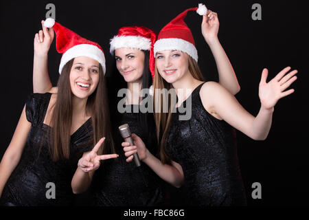 Gruppe von drei positiv, glücklich lächelnde schöne Mädchen Sänger posiert in roten Weihnachtsmann Mützen mit Mikrofon, schwarz backgr Stockfoto