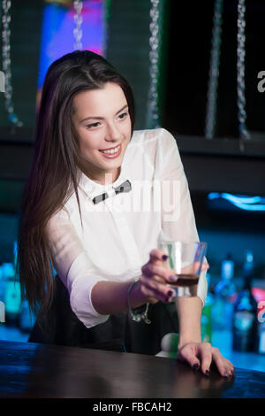 Schöne fröhliche Brünette Barkeeper Mädchen in weißem Hemd und schwarzer Fliege, das Alkohol trinken zu Disco Bar serviert, mit gla Stockfoto