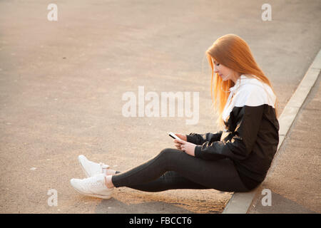 Sportliches Girl Sportswear mit einem Gadget, Handy oder Player, sitzt auf dem Bürgersteig im warmen Morgenlicht, Textfreiraum Stockfoto