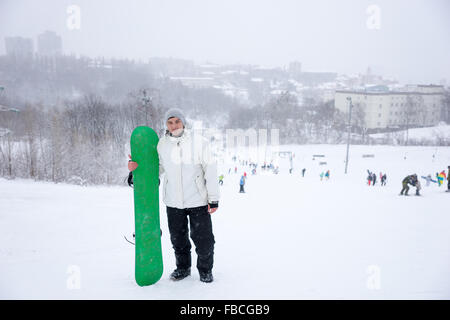 Junger Mann mit einem bunten grün Snowboard stehen auf einem verschneiten Berg posiert für die Kamera mit Massen von Snowboardern, die in der Ferne sichtbar. Stockfoto