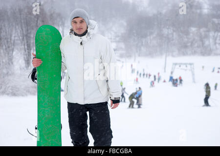 Sportlicher junger Mann hält ein helles Grün Snowboard auf einem verschneiten Berg laufen in einem Skigebiet posiert für die Kamera mit einem Lächeln Stockfoto