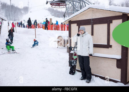 Junge Snowboarder mit Kaffee aufwärmen, steht er in der Nähe eine Skipiste in einem Resort mit Touristen als eine Unschärfe im Hintergrund sichtbar. Stockfoto