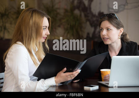 Zwei junge kaukasischen Frauen sitzen am Tisch im Café neben Laptop, Diskussion, blondes Mädchen hält Dokumentordner öffnen Stockfoto