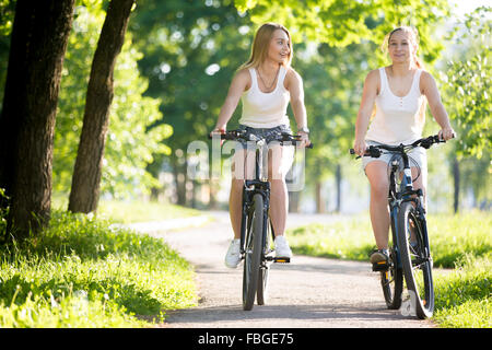 Zwei glücklich lächelnde junge Frauen Freundinnen tragen Jeans Hosen Fahrrad auf der Straße an sonnigen Sommertag genießen es, Bran Stockfoto