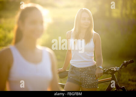 Zwei attraktive Freundinnen tragen lässige weiße Tank-Tops und Jeans-Shorts mit Fahrrädern im Natur Park auf helle Sonne stehend Stockfoto