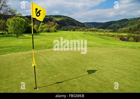 Gelbe Flagge in das Loch auf dem Grün Golfplatz, Fairway neben einem Teich, bewaldeten Bergen im Hintergrund Stockfoto