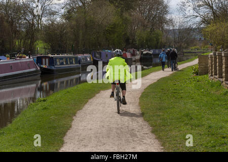 Vorbeifahrenden vertäut schmale Boote, ein Radfahrer und Wanderer Freizeit verbringen, Ausübung auf dem Treidelpfad - Leeds-Liverpool Kanal, Bingley, West Yorkshire, Großbritannien. Stockfoto