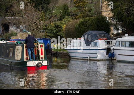 Mann steuert ein Narrowboat am Leeds-Liverpool-Kanal entlang, vorbei an moderner Kreuzfahrtschiffe vor Anker in der Nähe - Bingley, West Yorkshire, England, GB, UK. Stockfoto