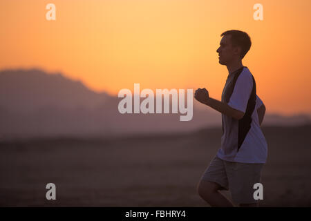Sportlicher junger Mann tun Lauftraining auf der Dämmerung vor Sonnenaufgang in Dessert mit Bergkette beleuchtet durch die aufgehende Sonne auf die backg Stockfoto