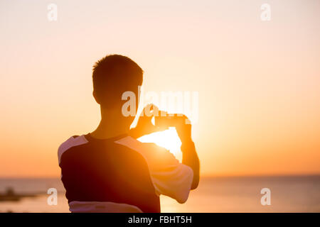Junger Mann halten Handy, unter Bild von Sonnenauf- oder Sonnenuntergang in malerischen Landschaften, Sonne, die durch seine Hände zurück Stockfoto