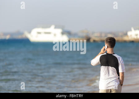 Junger attraktiver Mann hält Smartphone, app verwenden, machen Anruf telefonieren mit Handy während des Gehens im Sea Shore, weiße yac Stockfoto