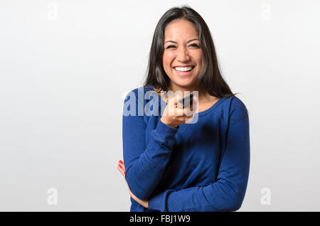 Einzelne Brünette Frau Lachen in blau, die Fernbedienung in die Kamera zeigen, als ob vor dem Fernseher Stockfoto