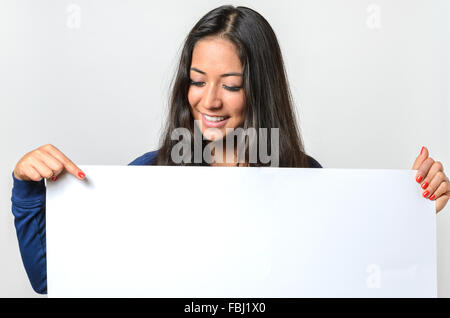 Attraktive junge Frau, die auf ein leeres weißes Schild mit textfreiraum für Ihren Text, den sie vor ihr hält lächelnd Stockfoto