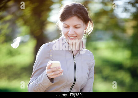Porträt von glücklich lächelnde kaukasischen Frau im Park auf der Bank, Standortwahl mit Handy, messaging Stockfoto