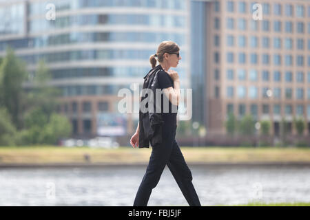 Junge Geschäftsfrau schnelles gehen in der Stadt neben River Bank und Bürogebäuden, halten ihre Jacke auf der Schulter, Profil vi Stockfoto