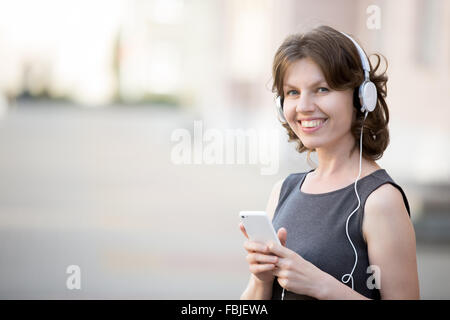 Porträt der glückliche Frau in weißen Kopfhörer Handy in den Händen auf Straße halten, im Sommer mit app, Musik hören, Freund Stockfoto