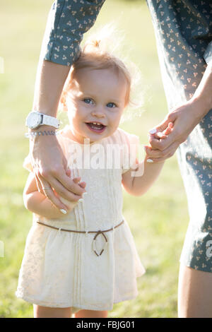 Junge Mutter, die Lehre um zu gehen fröhlich entzückende blonde Kleinkind Mädchen im weißen Kleid, Mutter ihre kleine Tochter Händchenhalten, suppor Stockfoto