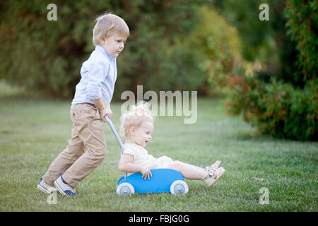 Porträt von zwei glücklichen niedlichen kleinen Geschwister spielen im Freien. Bruder mit seiner Schwester Kleinkind in Spielzeug Kinderwagen, Wandern Zusam Stockfoto