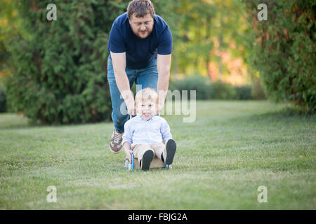 Vater und Sohn spielen, gemeinsam Spaß haben im Park im Sommer. Vater mit seinem Kind glücklich und aufgeregt in Spielzeug Kinderwagen mit Geschwindigkeit, Stockfoto