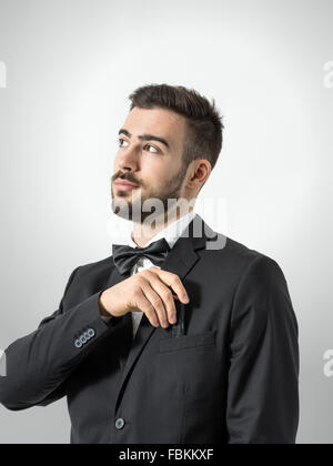 Junge Bräutigam im Smoking mit Fliege Umsetzung Binderücken in seinem Anzug Tasche. Entsättigt Porträt über graue Studio-Hintergrund Stockfoto