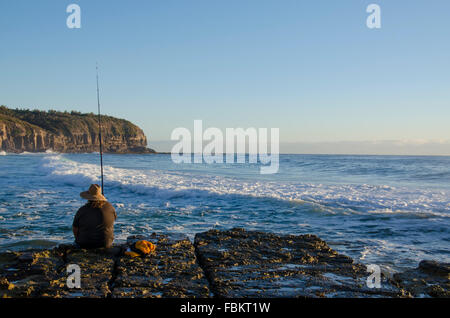 Eine kaukasische Person fischt in der Morgendämmerung am Rand einer felsigen Plattform, während Wellen hereinrollen und die Sonne an einem Strand von Sydney aufgeht Stockfoto