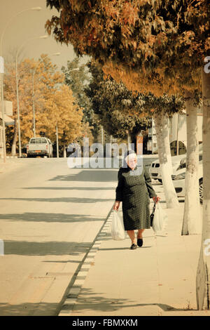 Alte Dame in schwarz in Zypern zu Fuß entlang der Straße mit Einkaufstüten in Bäumen gesäumten Allee Stockfoto