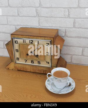 Hölzerne Mantel Vintage Art-déco-Uhr mit alten weiße Ziegel Wand Effekt Hintergrund und China Teetasse und mit Tee gefüllt Stockfoto