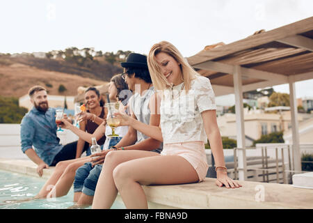 Im Freien Schuss glückliche junge Menschen feiern. Junge Männer und Frauen sitzen am Rand des Schwimmbades mit Füßen im Wasser Stockfoto