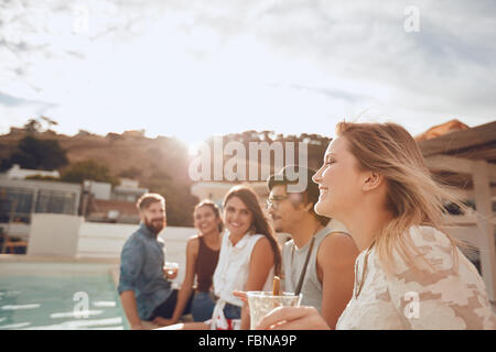 Gruppe von Freunden sitzen in einer Reihe am Rande des Schwimmbad und genießen Partei. Fröhliche junge Menschen mit einer Party auf dem Dach Stockfoto