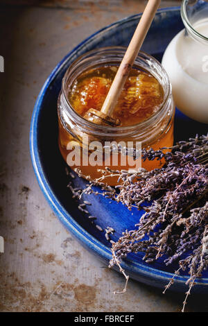 Offenen Glas flüssiger Honig mit Waben und Honig Wagen im Inneren, Glaskrug mit Milch und Haufen von trockenen Lavendel in großen blauen c Stockfoto