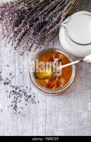 Offenen Glas flüssiger Honig mit Waben und silbernen Löffel im Glaskrug mit Milch und Haufen von trockenen Lavendel über weiß wo Stockfoto