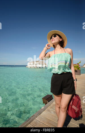 schlanke Frau mit Hut und Sonnenbrille posting auf Holzbrücke über dem kristallklaren Meerwasser auf den Malediven an einem sonnigen Tag Stockfoto