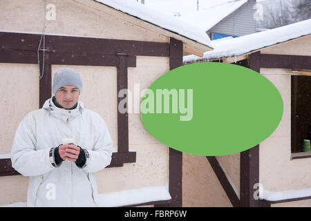 Ruhig Jüngling im Mantel mit Heißgetränk neben leeren grünen Schild mit textfreiraum auf einer Skihütte Stand Stockfoto