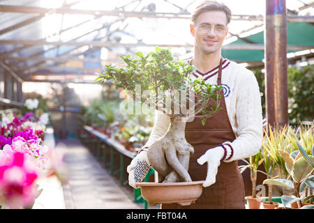 Porträt von attraktiven glücklich männlichen Gärtner mit Schürze und Handschuhe stehen und hält Bonsai-Baum im Topf im Garten-center Stockfoto