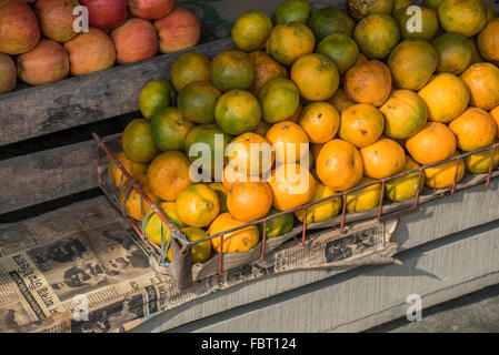 Lebendige frische Orangen und Äpfel zum Verkauf auf einer Holzkiste mit Zeitung in warmem Abendlicht. Markt im Fährhafen Majuli Island. Assam, Indien Stockfoto