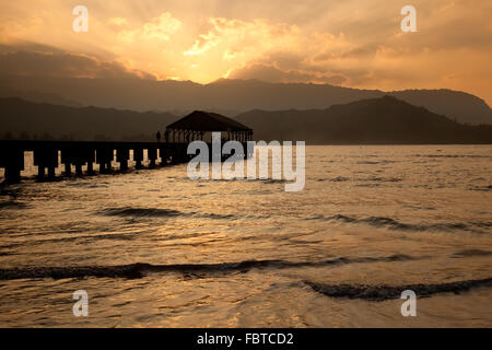 Sonnenuntergang hinter Na Pali Bergen mit einer Silhouette des Hanalei Pier im Vordergrund und die Sonne reflektiert auf dem Ozean Stockfoto