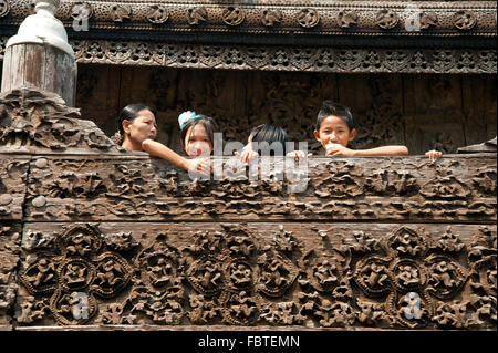 Die burmesische Kinder Peer über die hölzerne Wände vor dem Teakholz geschnitzt Wand des Shwenandaw Kyaung Pagode in Mandalay, Myanmar Stockfoto