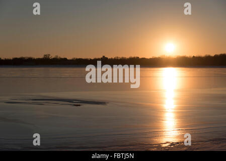 Die Sonne geht über der gefrorenen See, ohne Schneedecke, so dass die Sonne hell aus der IT. spiegelt. Stockfoto